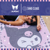 A person lying on a large Kuromi-themed beach towel or blanket outdoors, with a branded top banner showing Kuromi and Sand Cloud logos. The product is light pink with a large gray and white Kuromi face graphic and small star accents.