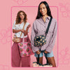 Two women modeling patterned tote and crossbody bags against a pink floral background. The woman in front wears a pink-and-white check shirt and denim shorts, holding a black crossbody bag with a floral print.