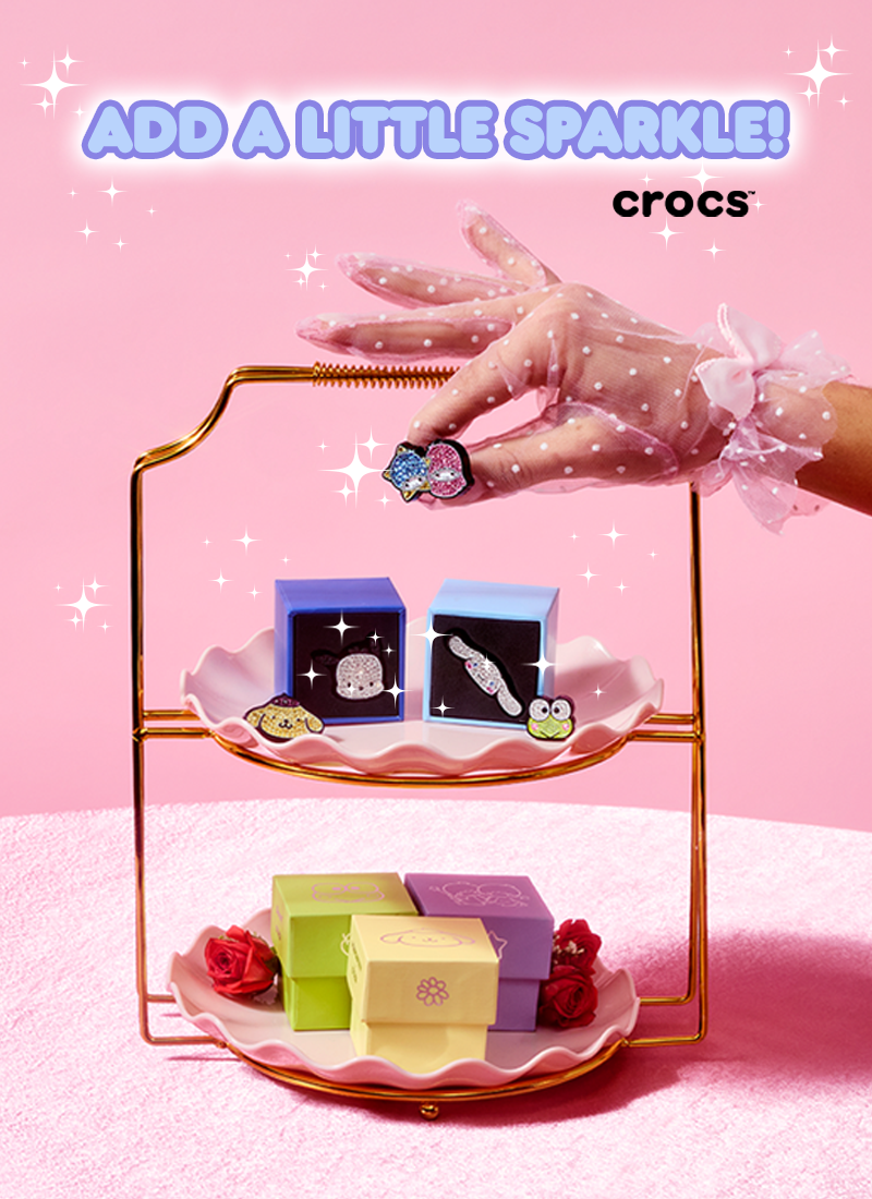 Three-tiered tray with small boxes and a hand wearing a polka dot glove with a bow, on a pink background with 'crocs' branding.